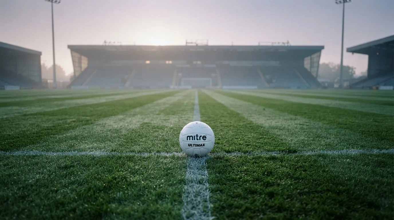 Ein Fußball liegt auf dem Mittelkreis eines leeren Rasenplatzes am frühen Morgen, im Hintergrund eine ferne Tribüne im Dunst.
