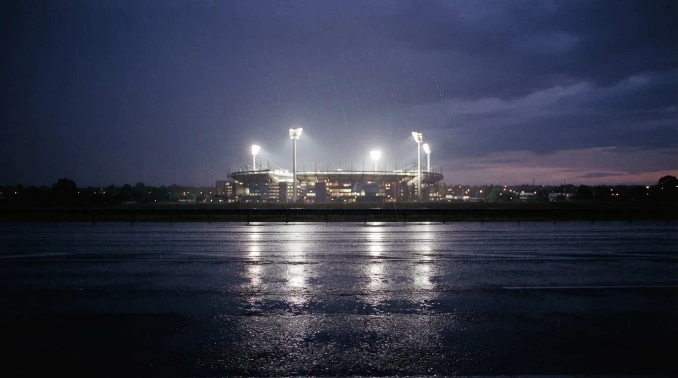 Ein Stadion unter Flutlicht aus großer Entfernung gesehen, davor eine regennasse Straße in der Dämmerung.