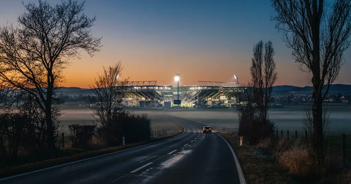 Ein Stadion unter Flutlicht aus großer Entfernung gesehen, davor eine dunkle Landstraße und einzelne Bäume in der Abenddämmerung.