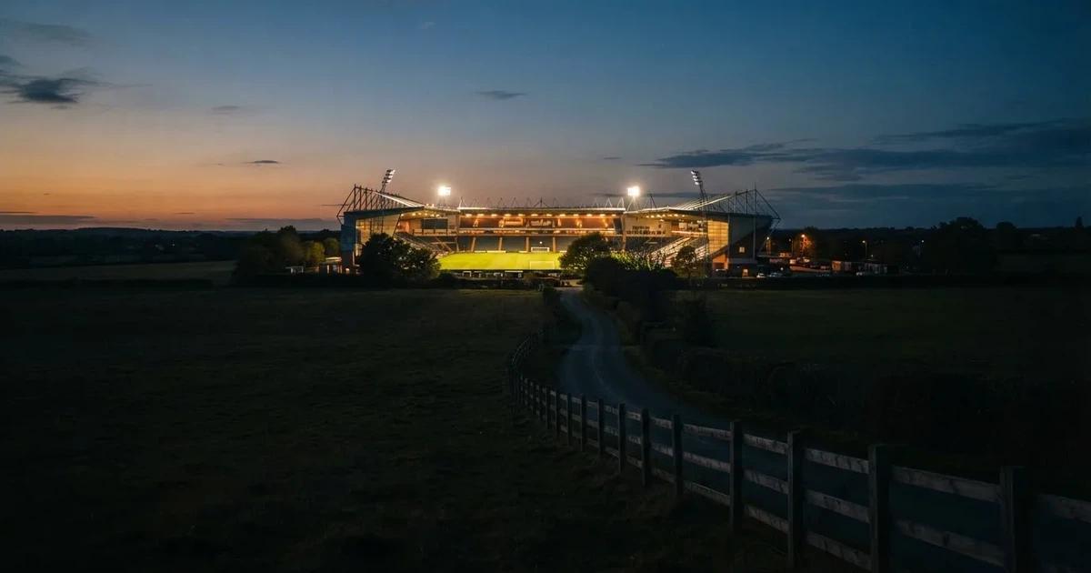 Ein Fußballstadion unter Flutlicht aus großer Entfernung gesehen, davor Felder und eine ruhige Straße in der Dämmerung.