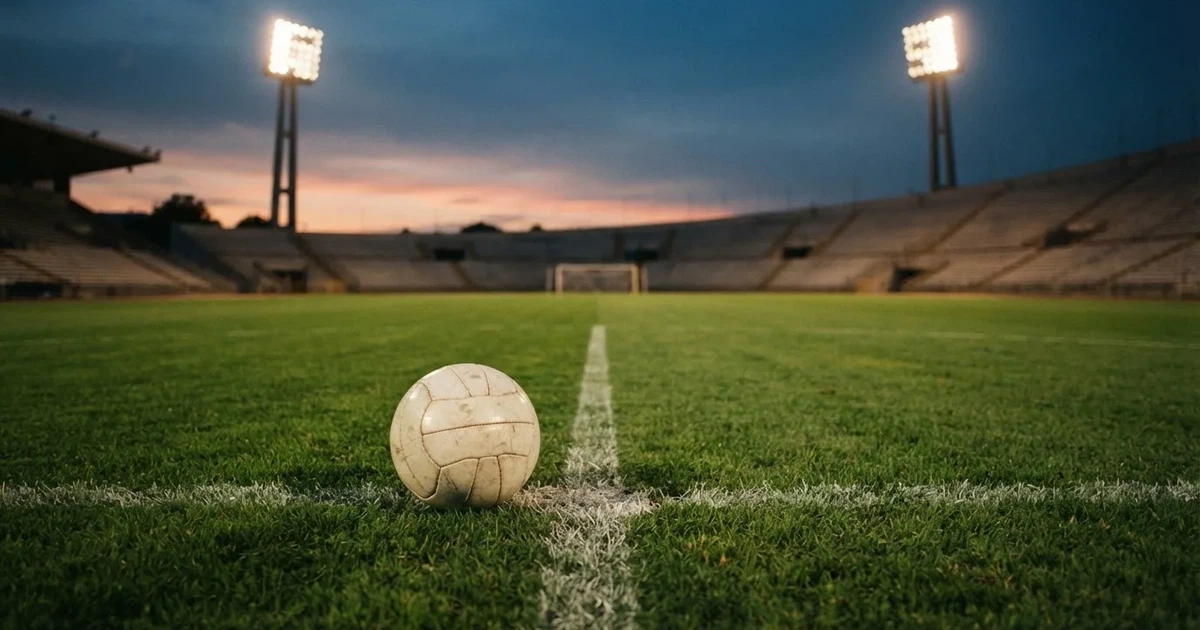 Ein Fußball liegt auf dem Anstoßpunkt eines Rasenplatzes in der Dämmerung, in der Ferne leuchten Flutlichter, die Ränge sind leer.